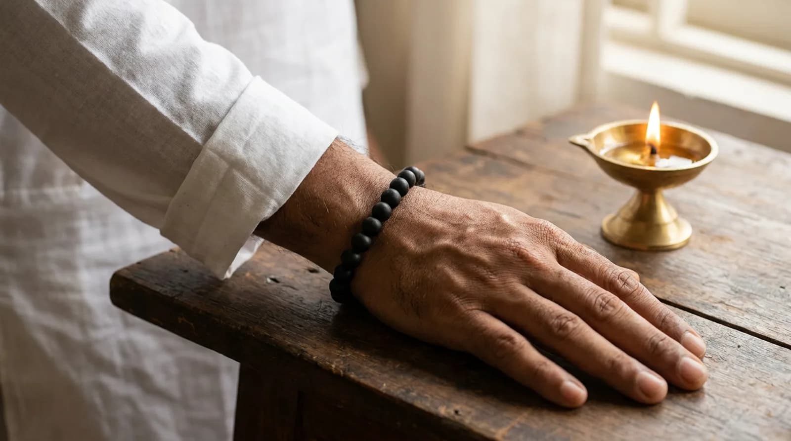 A man's wrist wearing a matte black karungali ebony wood bead bracelet beside a brass diya